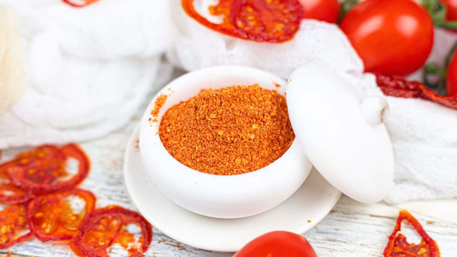 Ground tomato powder in a white bowl surrounded by dried and fresh tomatoes on a rustic wooden table.