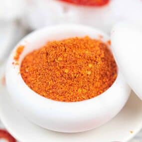 A close-up image of a bowl of vibrant orange-red tomato powder, placed on a white surface.
