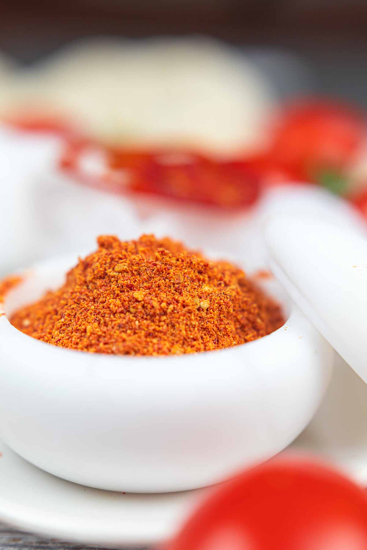 Ground tomato powder in a white ceramic bowl, closely focused, with blurred sliced red tomatoes in the background.