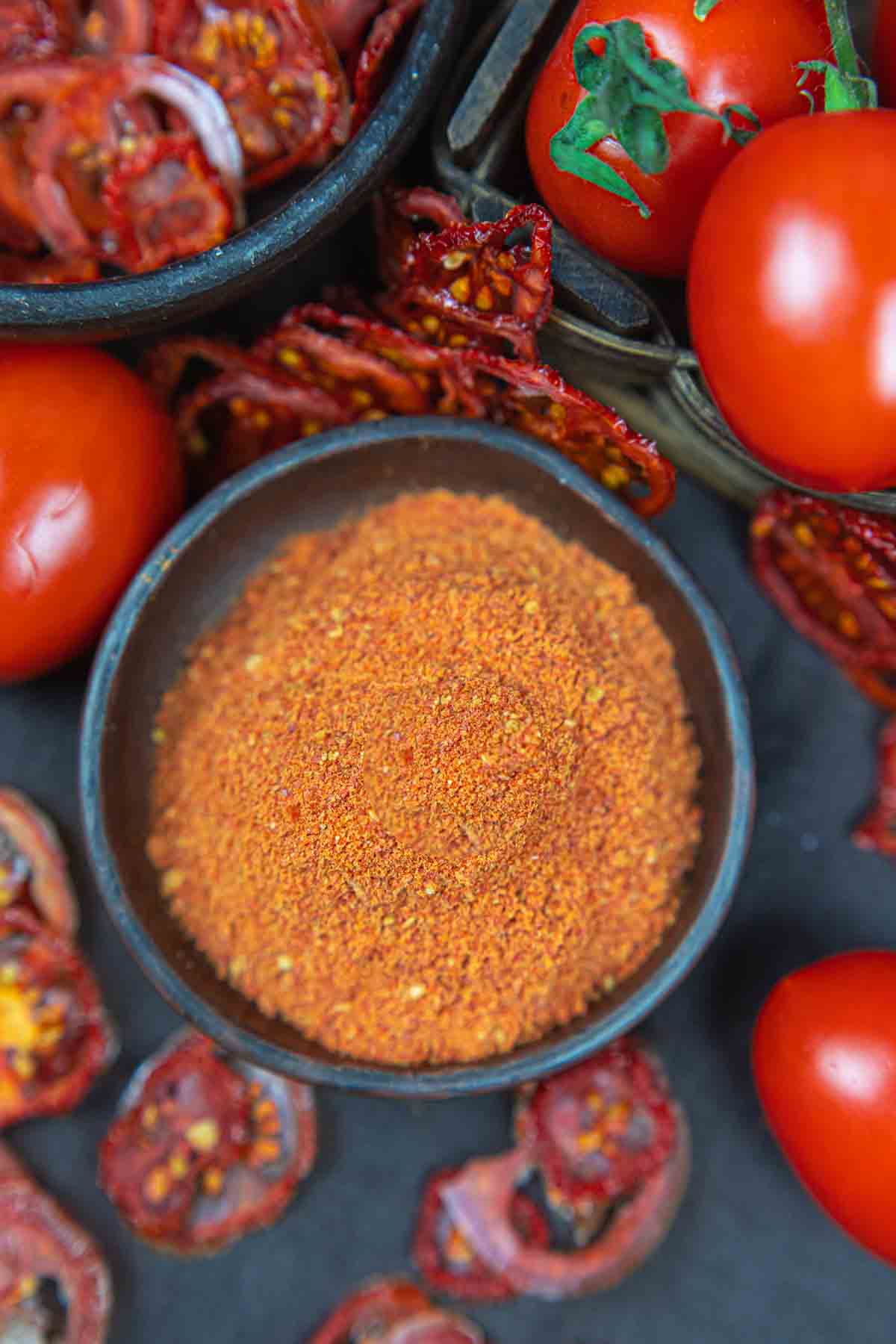 A close-up of a bowl of tomato powder surrounded by fresh tomatoes on a dark surface.