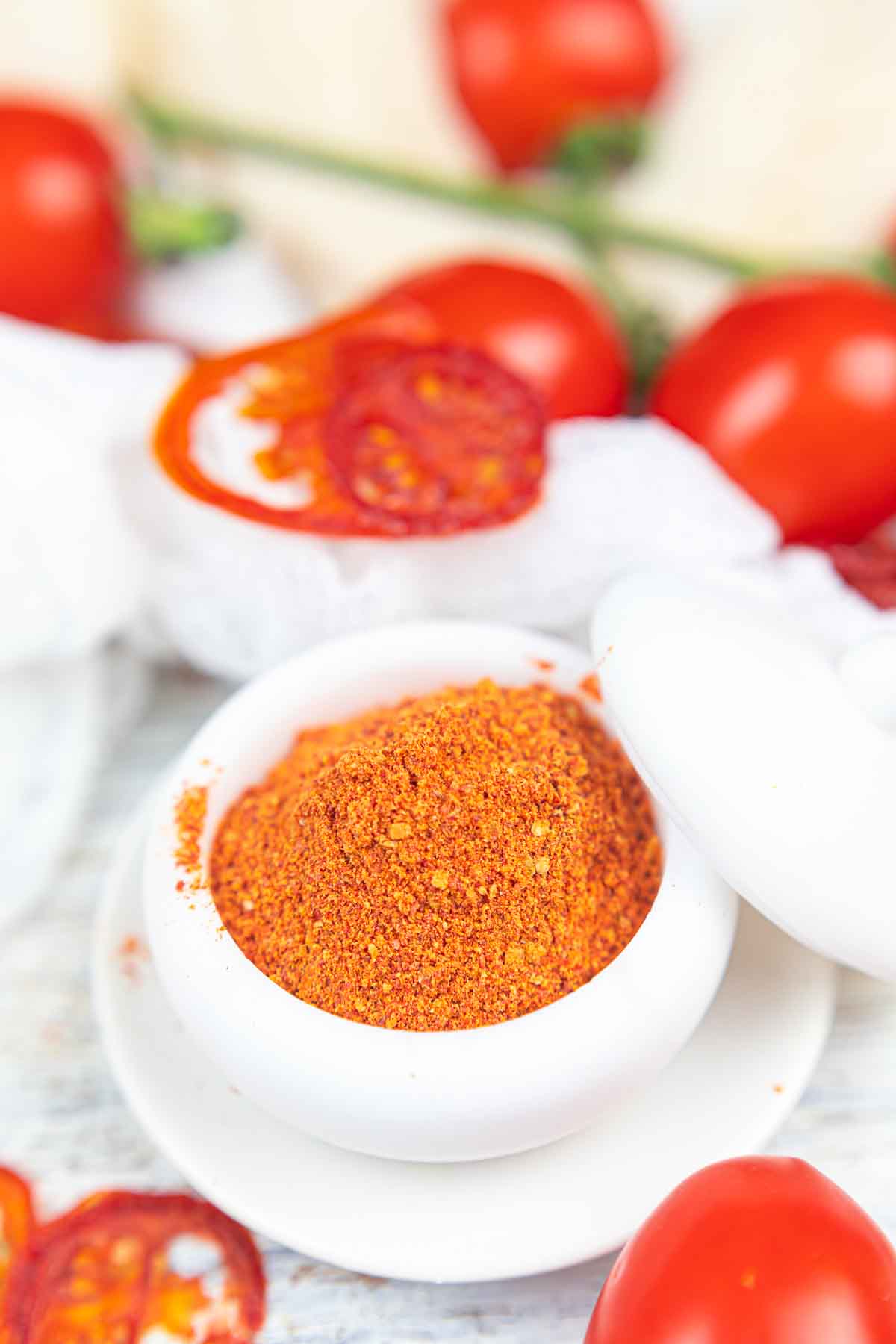 A small white bowl filled with tomato powder, surrounded by fresh whole and sliced tomatoes on a wooden surface.