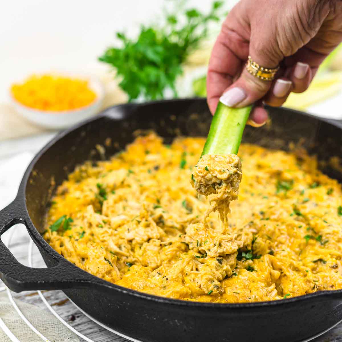 A hand dips a cucumber stick into a skillet of buffalo chicken dip.