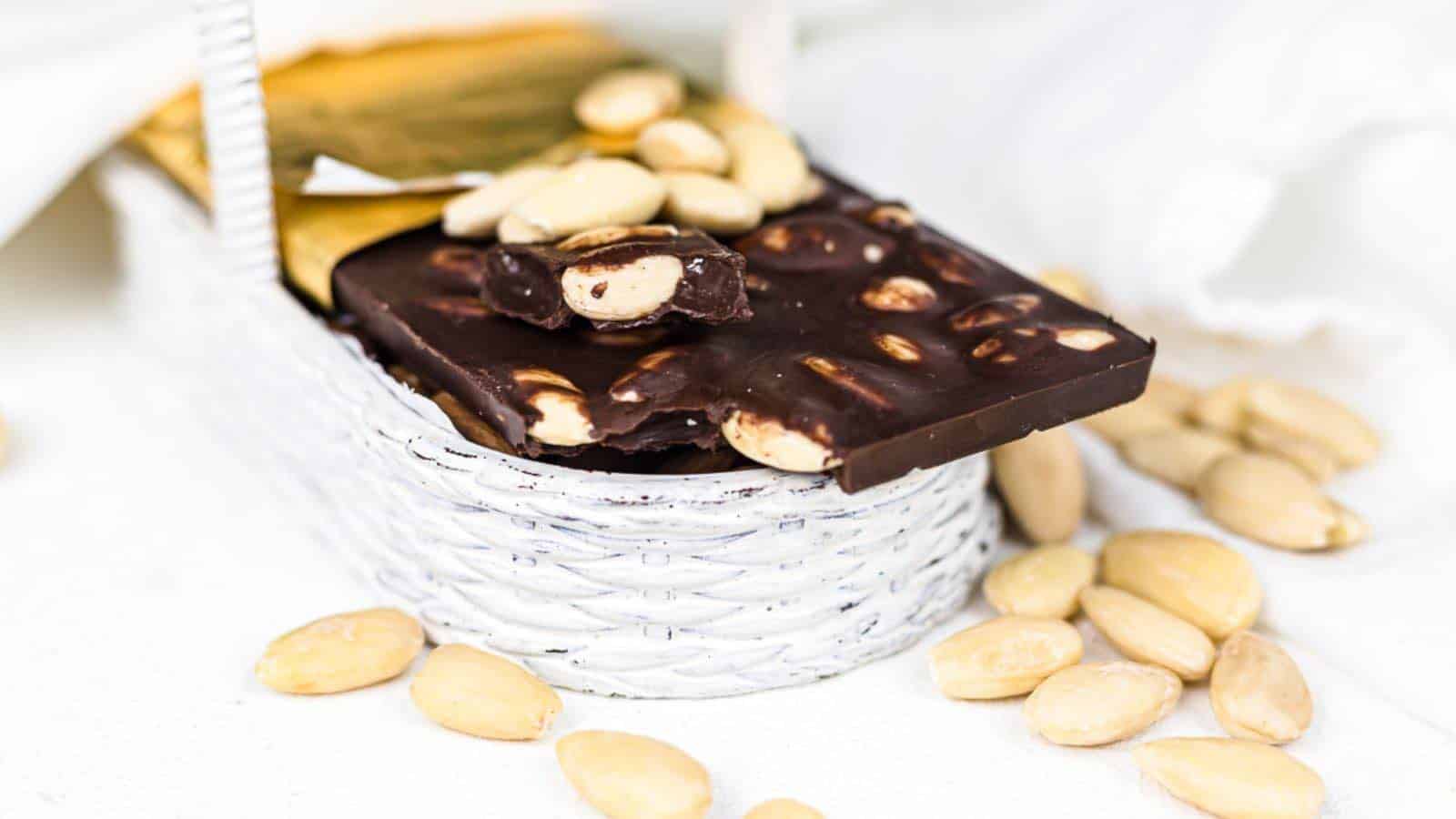 A bar of dark chocolate with nuts inside a small white woven basket, surrounded by scattered almonds on a white surface.