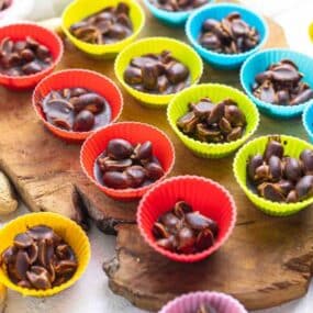 Colorful mini cupcake liners filled with chocolate and peanuts on a wooden board, surrounded by raw peanuts.