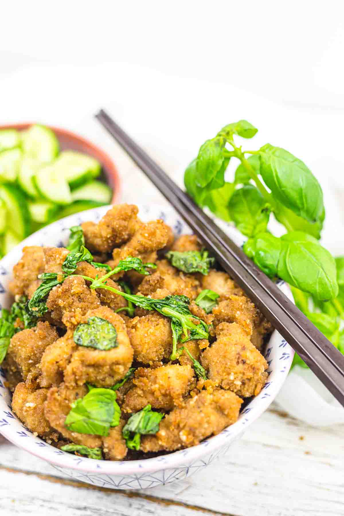 A bowl of breaded fried chicken with basil leaves, chopsticks on top, next to a bowl of sliced cucumbers.