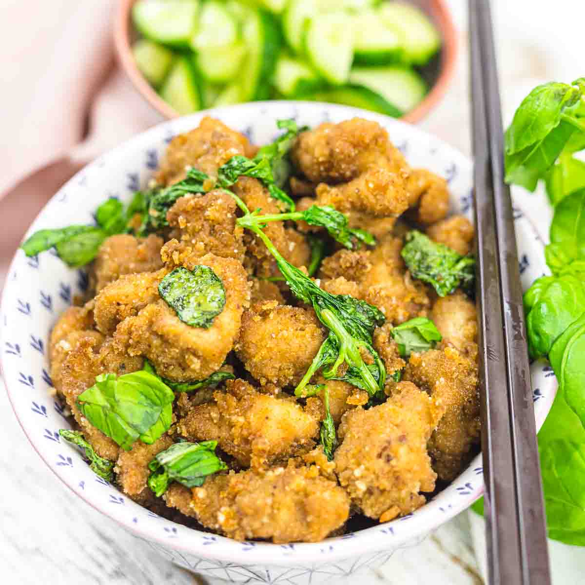 A bowl of crispy fried chicken topped with fried basil, served with a side of sliced cucumbers and fresh basil leaves.