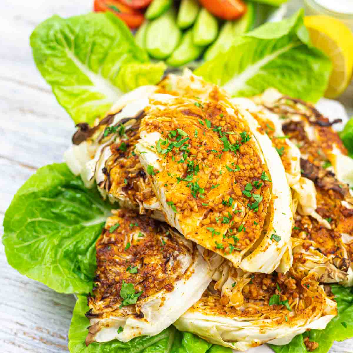 Grilled cabbage wedges garnished with chopped herbs, served on fresh lettuce leaves with a side of sliced cucumbers and tomatoes.