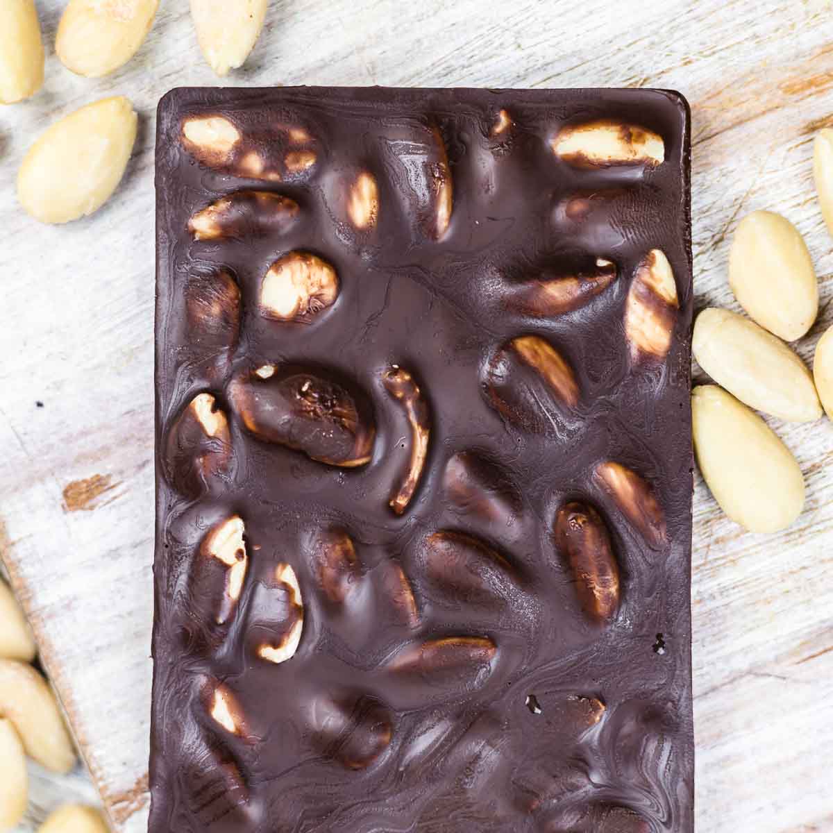 A close-up of a dark chocolate bar with whole almonds embedded in it, surrounded by a few loose almonds on a wooden surface.