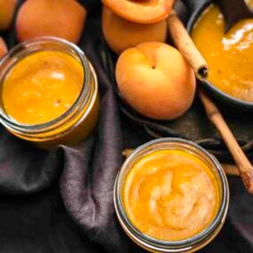 Two jars of apricot jam with whole peaches and a bowl of jam in the background, surrounded by cinnamon sticks.