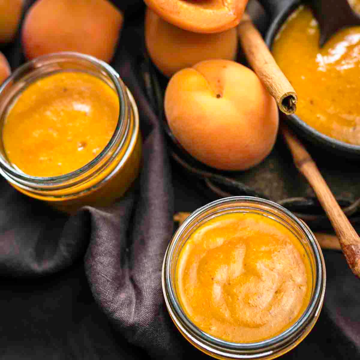 Two jars of apricot jam with whole peaches and a bowl of jam in the background, surrounded by cinnamon sticks.