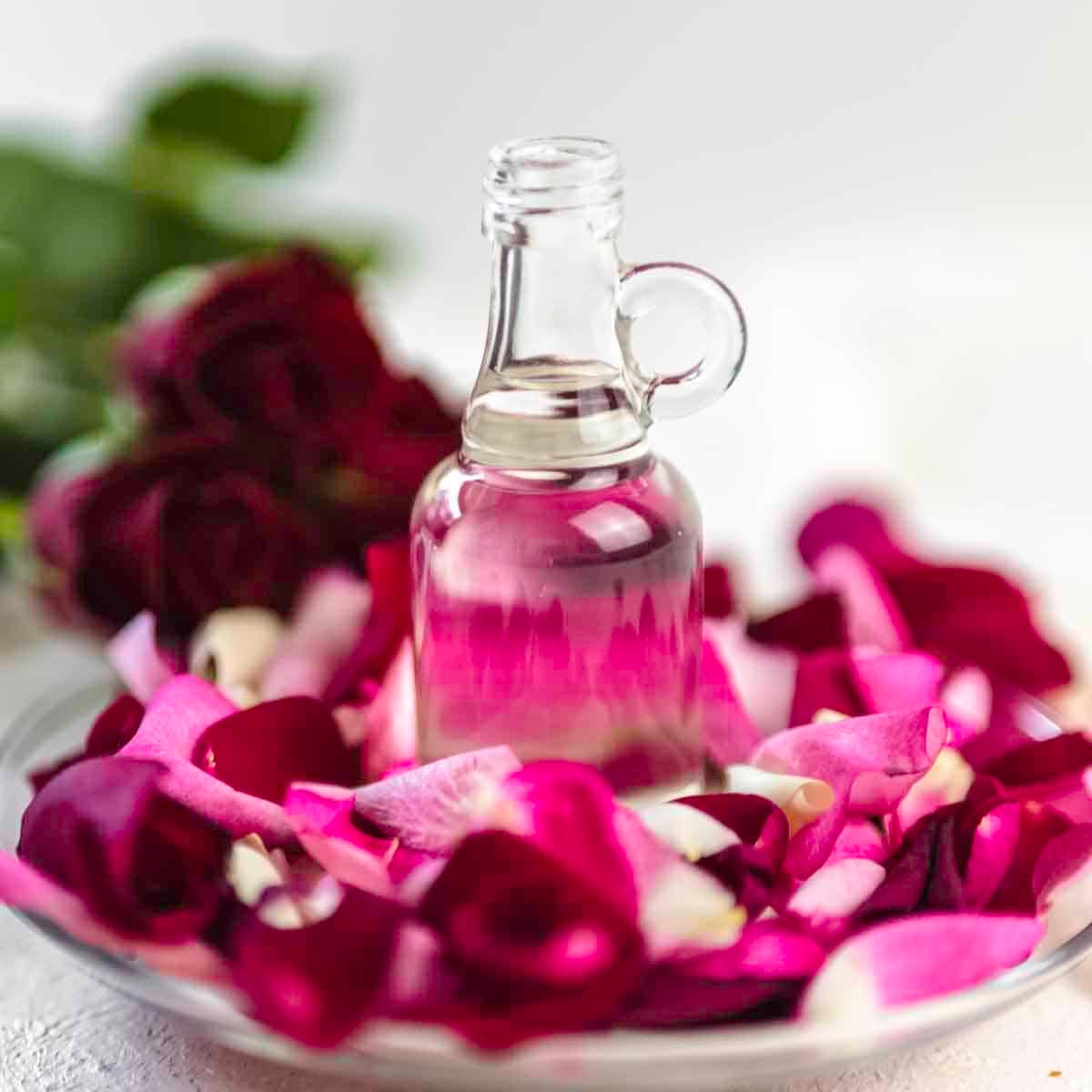 A clear glass bottle of rose water stands on a dish surrounded by red and pink rose petals.