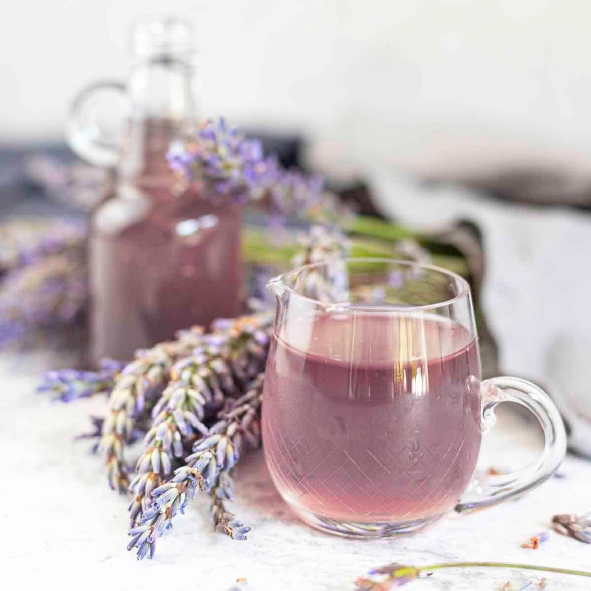 A glass pitcher filled with lavender syrup sits in front of a similarly filled bottle, accompanied by sprigs of fresh lavender flowers.
