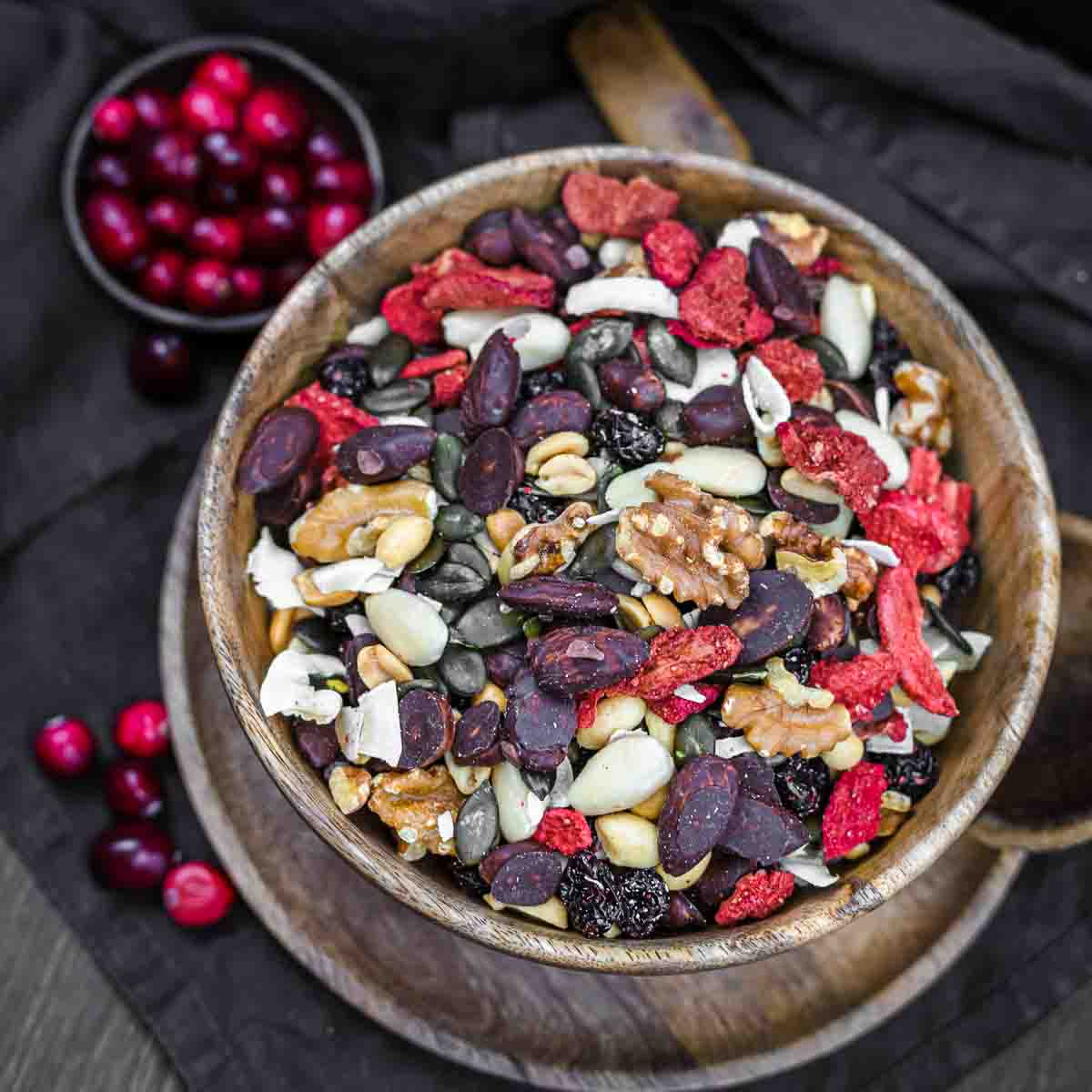 A wooden bowl filled with a trail mix of assorted nuts, seeds, and dried fruits on a dark cloth backdrop. A small dish of cranberries is in the background.