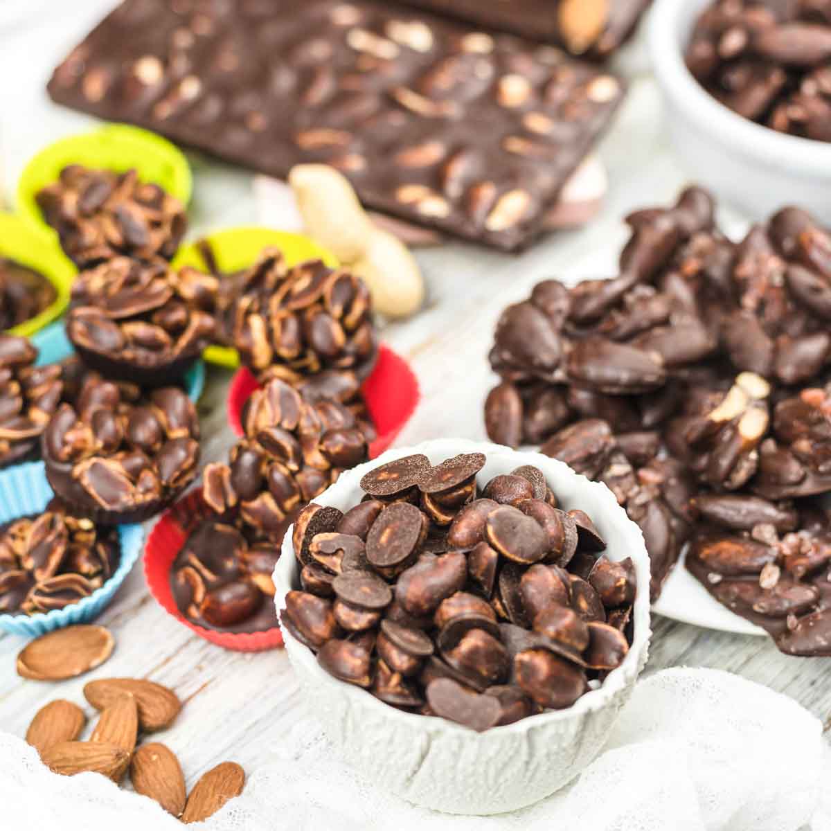 An assortment of chocolate-covered almonds and nuts displayed in bowls and colorful silicone cups on a light wooden surface.