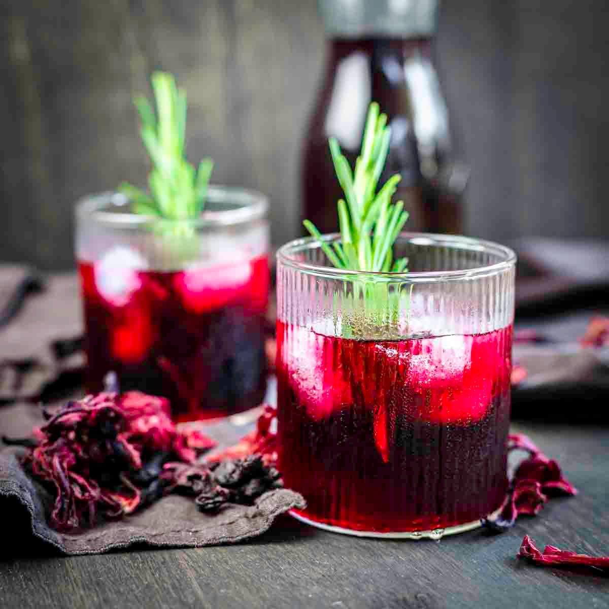 Two glasses filled with a dark red beverage, ice, and garnished with sprigs of rosemary. A bottle with more liquid is in the background, and dried flowers are scattered on a dark surface.