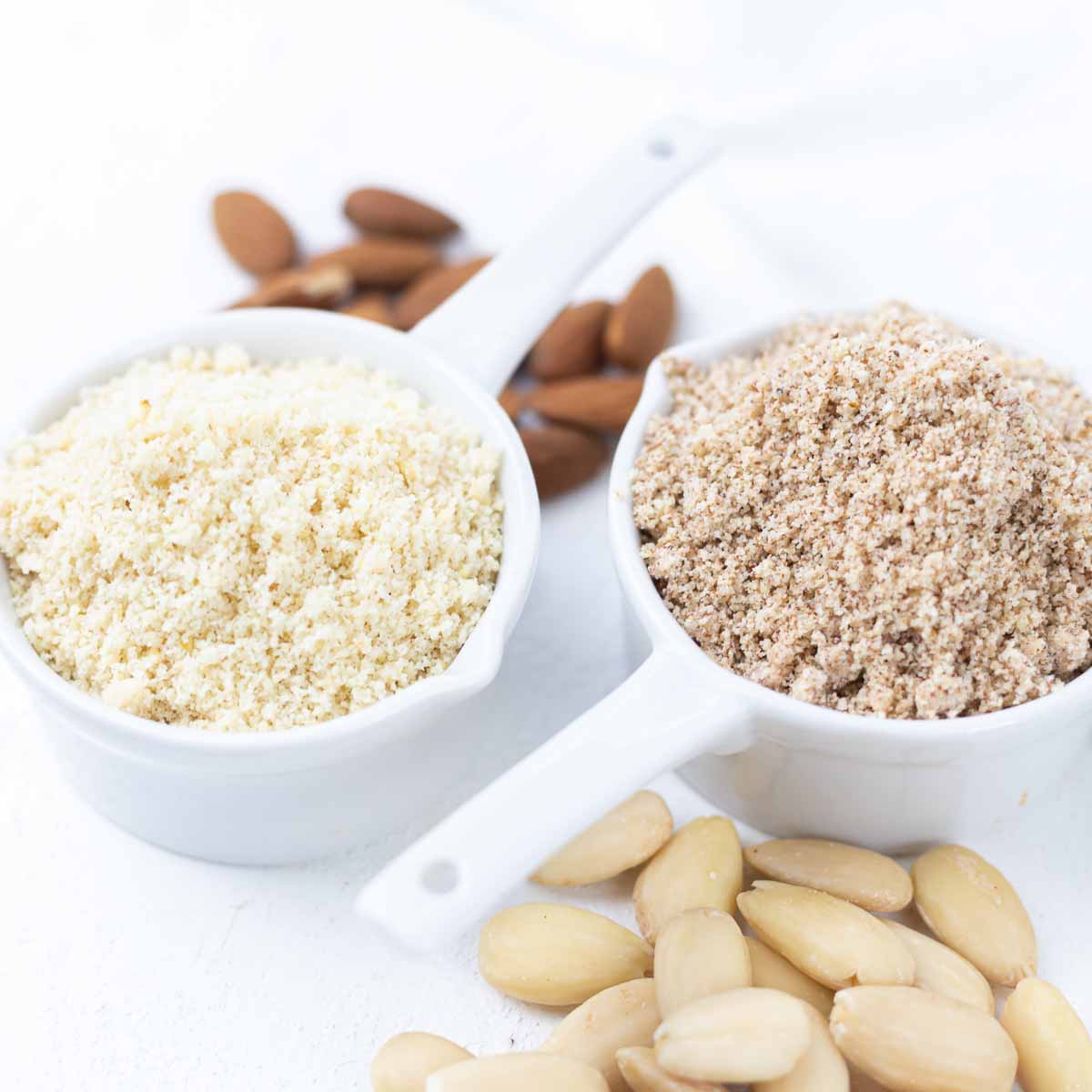 Two white measuring cups filled with almond flour and almond meal respectively. Whole almonds and blanched almonds are scattered around them on a white surface.