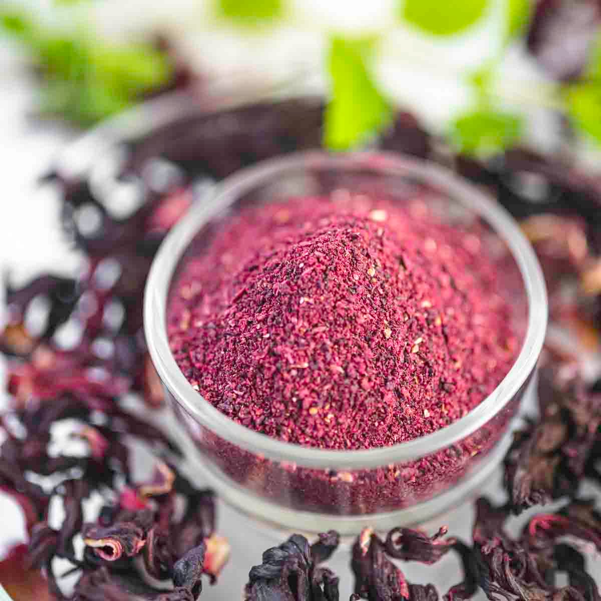 A small glass bowl filled with a fine, dark reddish-purple powder is surrounded by dried hibiscus petals.