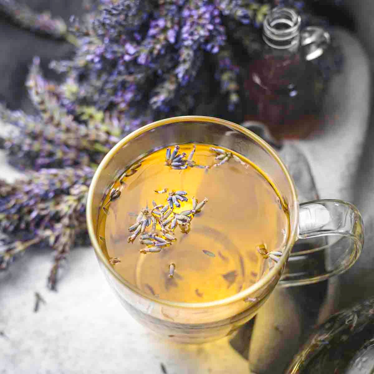 A glass cup filled with lavender tea garnished with lavender buds, with a small bottle and a bunch of fresh lavender flowers in the background.
