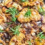 Cooked pieces of meat with caramelized onions and fresh rosemary sprigs in a pan.