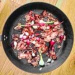 A frying pan on a wooden surface containing cooked meat pieces and small onions.
