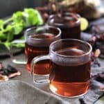 Three glass cups of hibiscus tea are arranged on a table with scattered dried hibiscus petals and a sprig of fresh mint.