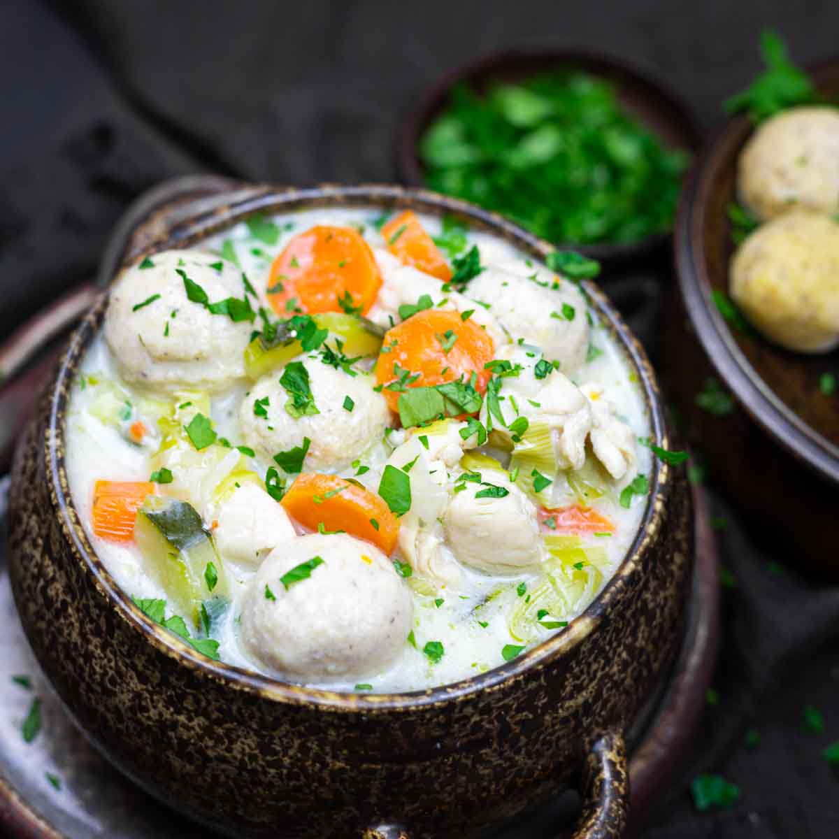 A bowl of creamy chicken and vegetable soup garnished with herbs, featuring chicken pieces, carrots, and celery, served on a dark background with a side dish of bread rolls.