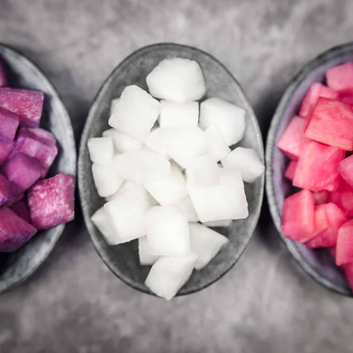 Three bowls containing diced vegetables: purple in the left bowl, white in the center, and pink in the right bowl, set against a gray background.
