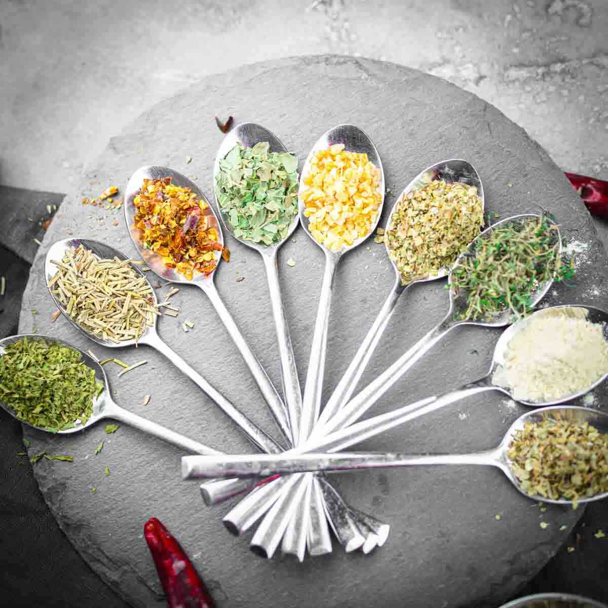 Nine spoons on a slate platter, each holding different dried herbs and spices, with two red chili peppers nearby.