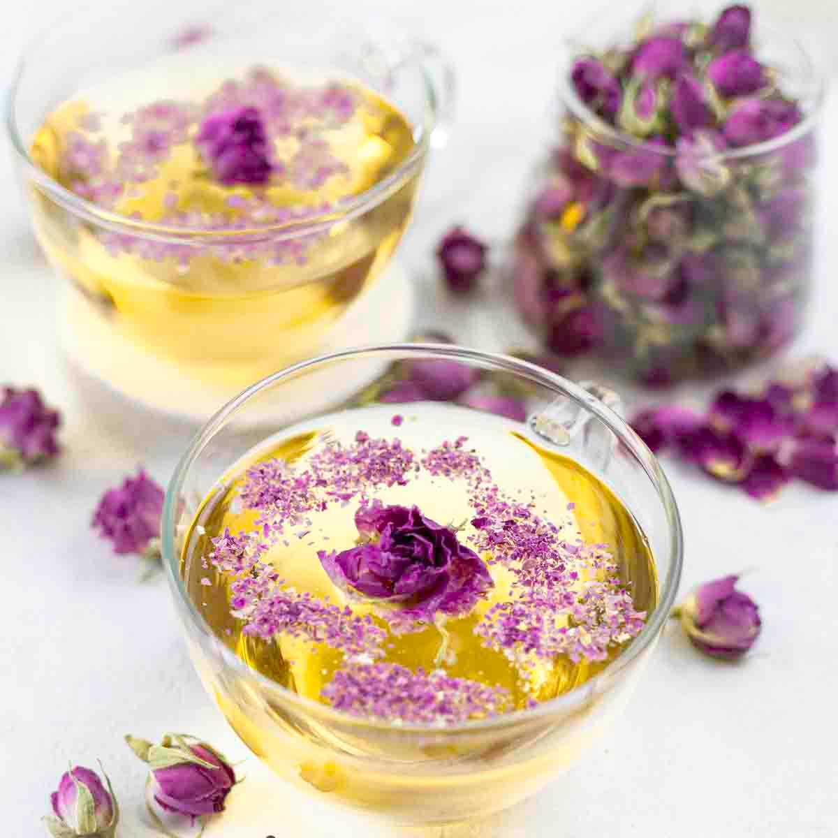 Two clear teacups filled with yellow liquid, garnished with purple flowers. A jar of similar flowers is in the background, with scattered flowers around.