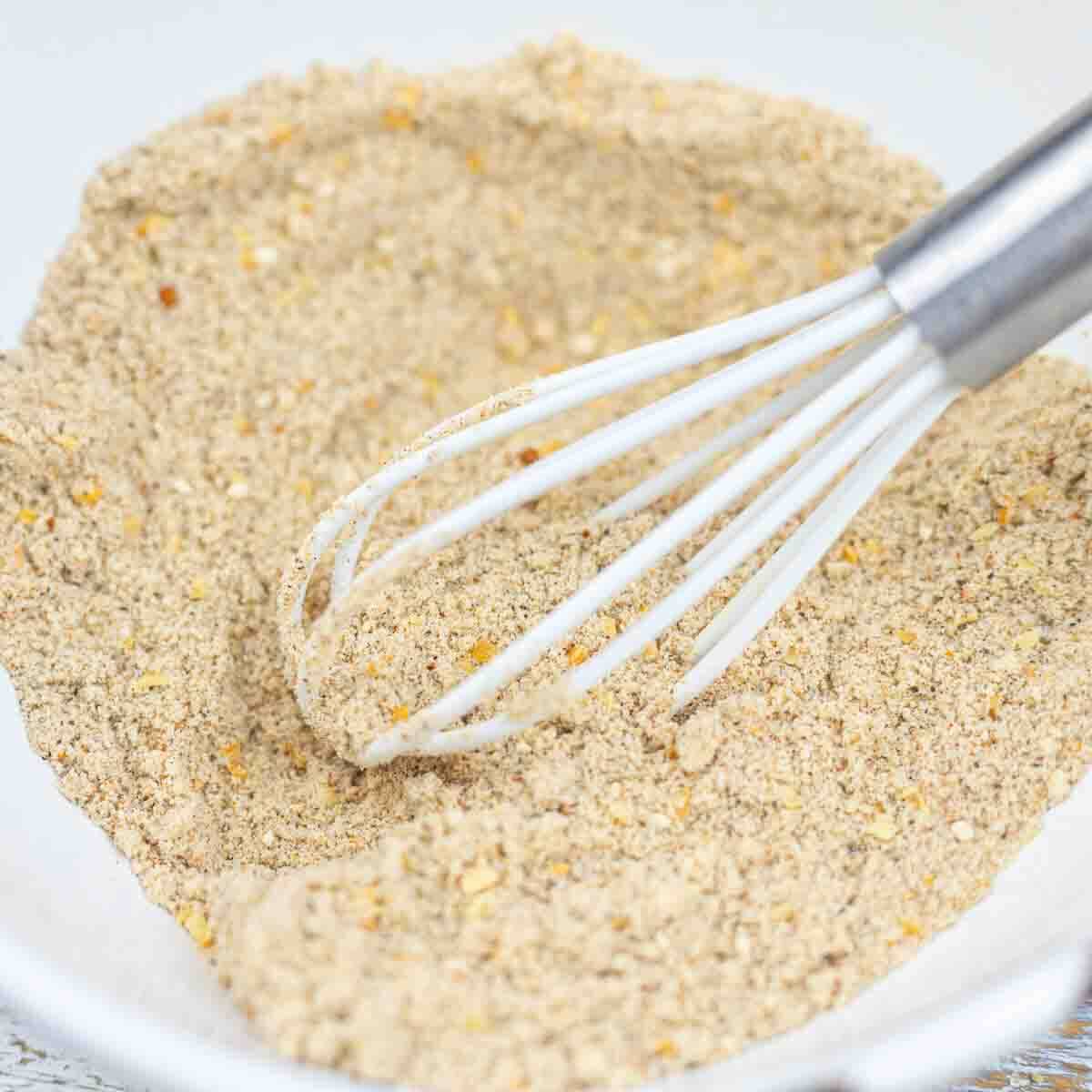A whisk rests in a white bowl containing a coarse, beige dry mixture.