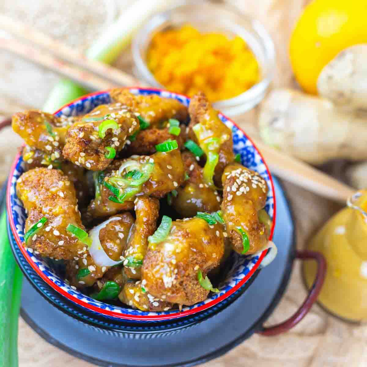 A bowl of fried chicken wings coated in a sticky glaze, garnished with sesame seeds and green onions. Yellow powder and fresh ingredients are in the background.