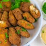 A bowl of crispy falafel garnished with herbs, accompanied by a dipping sauce and a small side of chopped green herbs.