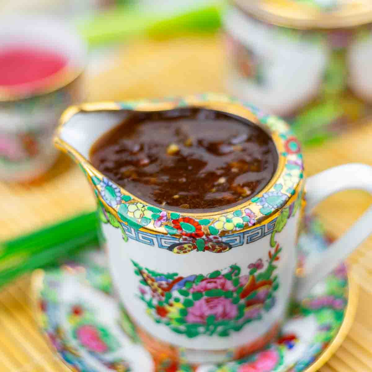 A decorative porcelain jug filled with a thick, dark sauce sits on a matching saucer. The set features colorful floral patterns and gold trim.