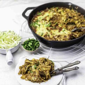 Cast iron skillet with baked pasta topped with cheese and herbs. A serving on a white plate with forks beside small bowls of shredded cabbage and chopped herbs, all on a wire rack.