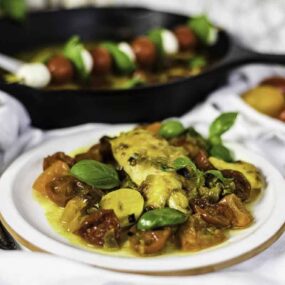 A plated dish featuring stuffed pasta with sauce and garnished with basil, accompanied by a black skillet with skewered cherry tomatoes and cheese in the background.