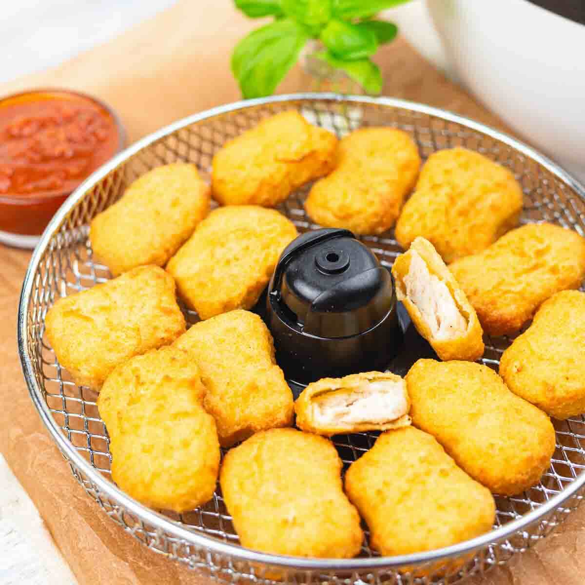 Golden chicken nuggets arranged in a circular basket with a dark central hub. A small bowl of red dipping sauce sits nearby on a wooden surface.