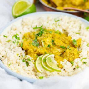 A white bowl with rice and yellow curry garnished with lime slices and herbs.