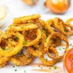 A pile of crispy onion rings garnished with chopped chives on a wooden board, accompanied by a small dish of dipping sauce.
