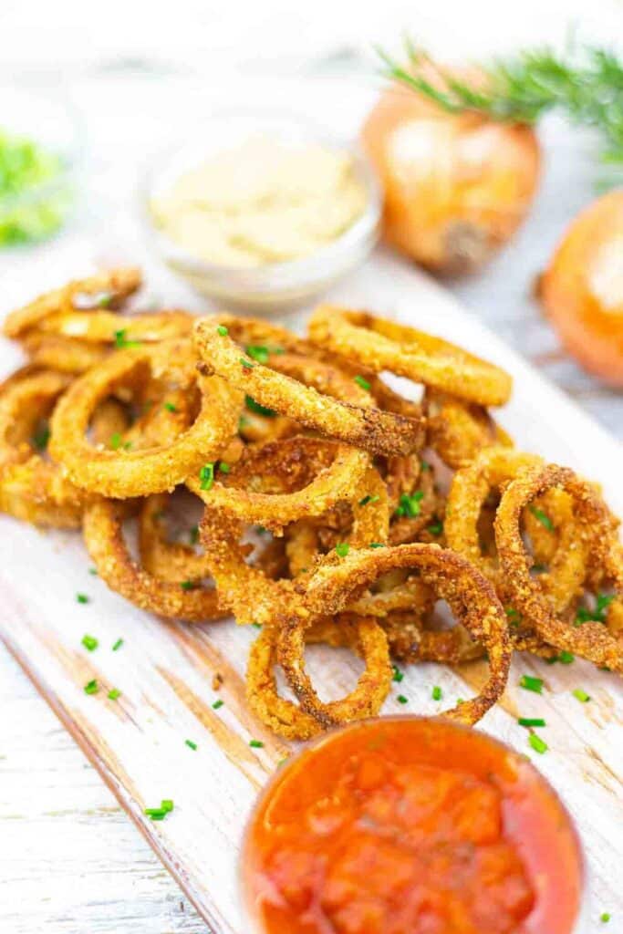 A stack of crispy onion rings on a wooden board, garnished with herbs. Accompanied by small bowls of dipping sauces, onions, and herbs in the background.
