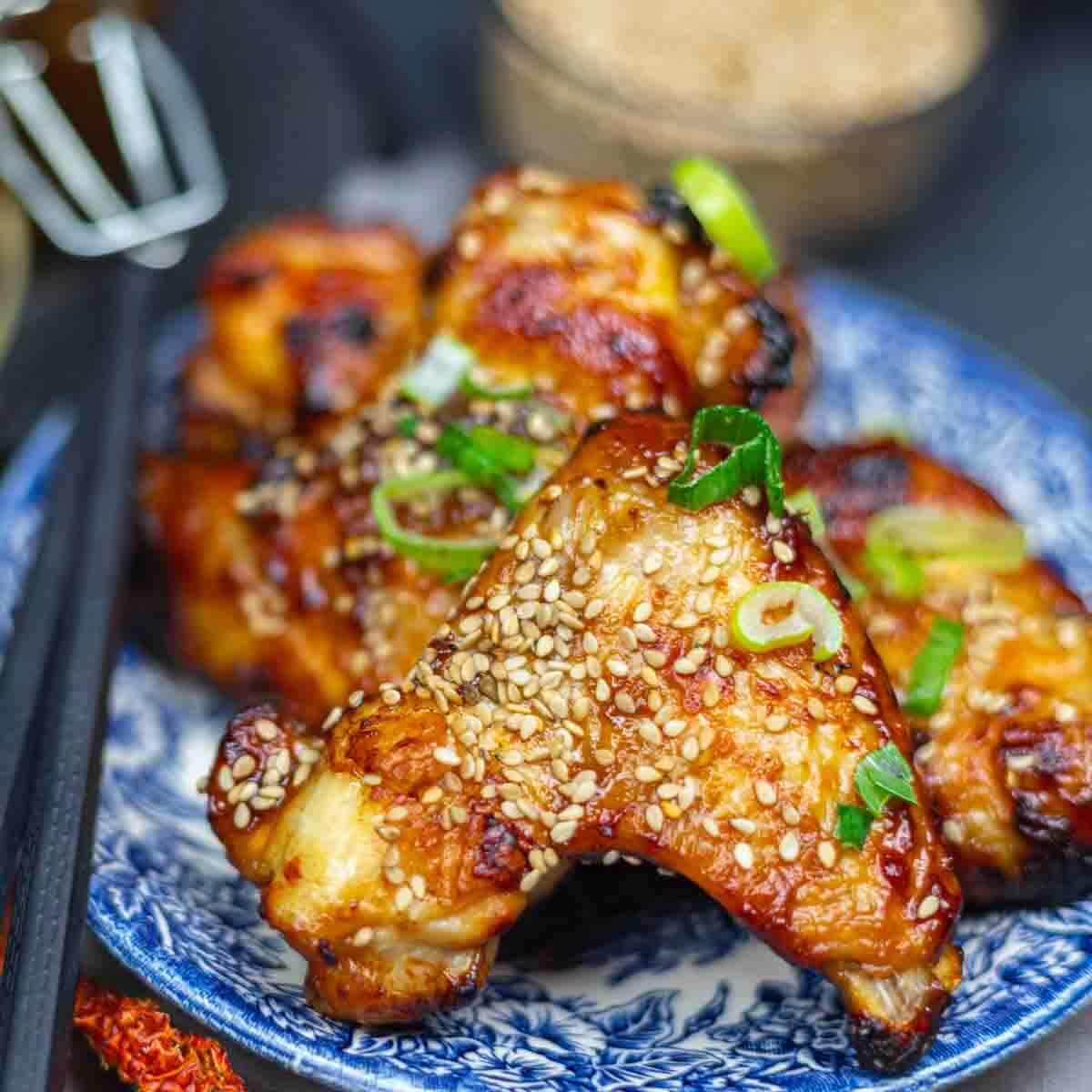 Plate of glazed chicken wings topped with sesame seeds and green onions, served on a blue patterned plate with chopsticks beside it.