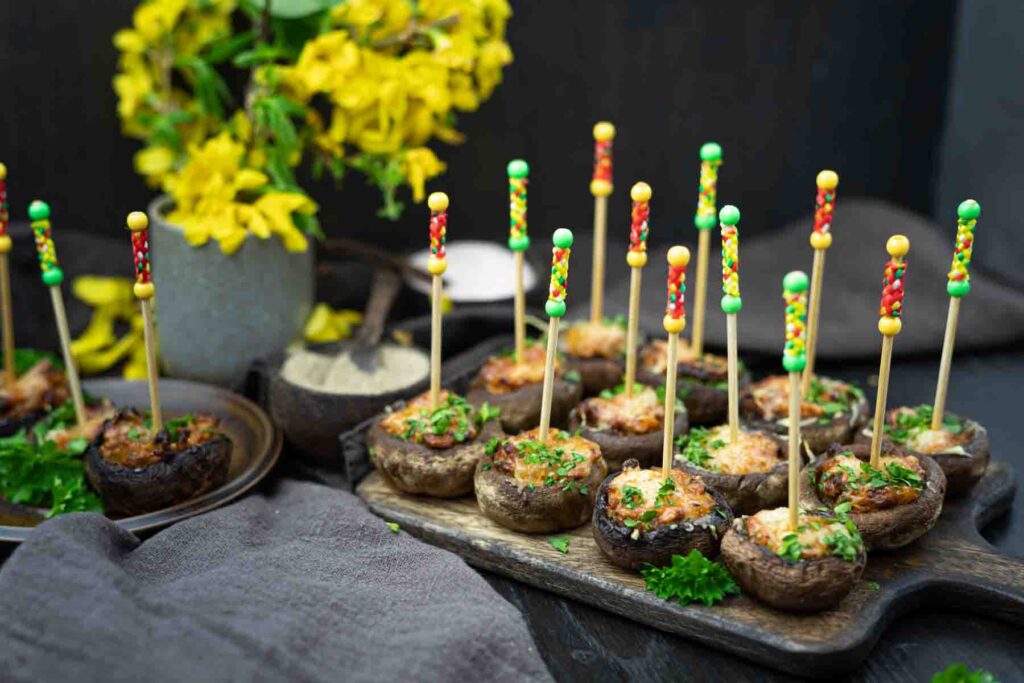 Stuffed mushrooms with colorful skewers on wooden trays, garnished with parsley. Yellow flowers in a vase are in the background, alongside bowls of seasoning.