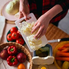 A person slices cabbage on a small cutting board over a grater, surrounded by tomatoes, apples, bell pepper, carrots, and other vegetables on a kitchen counter.