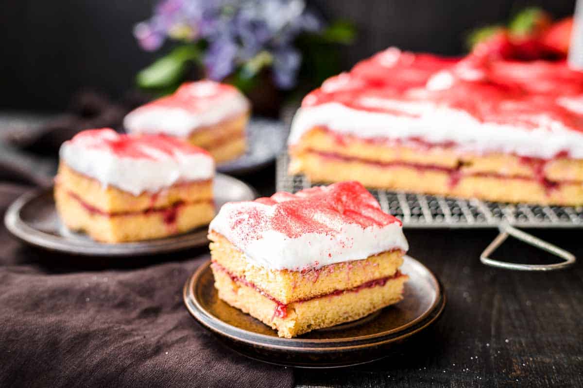 Slices of layered cake with white frosting and red topping, served on small plates. A larger portion is on a cooling rack in the background.