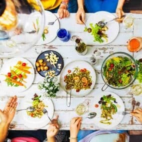 People sitting around a rustic table eating salads, roasted vegetables, cheese, and drinking beverages, with flowers as decoration.