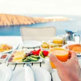 A hand holding a glass of orange juice over a breakfast table set with various dishes, overlooking a scenic, coastal landscape.