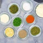 Nine small glass bowls contain various dried herbs and spices arranged on a gray surface, including salt, pepper, red paprika, chives, dill, and garlic.