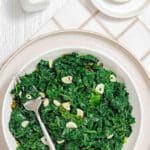 A plate of sautéed kale with sliced garlic, served with a fork on a white plate set on a checked napkin.