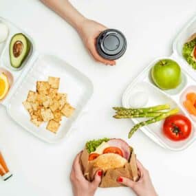 Three takeout containers with assorted foods, including sandwiches, vegetables, fruit, and crackers. Two hands hold sandwiches; a third hand holds a coffee cup. A bottle of sauce is nearby.