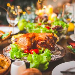 A roasted chicken is served on a platter with lettuce and tomatoes, surrounded by salads, pastries, empty plates, glasses, and festive decorations on a dining table.