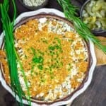 A baked casserole topped with golden breadcrumbs and chopped chives, surrounded by fresh rosemary, chives, and a bowl of sliced pickles.